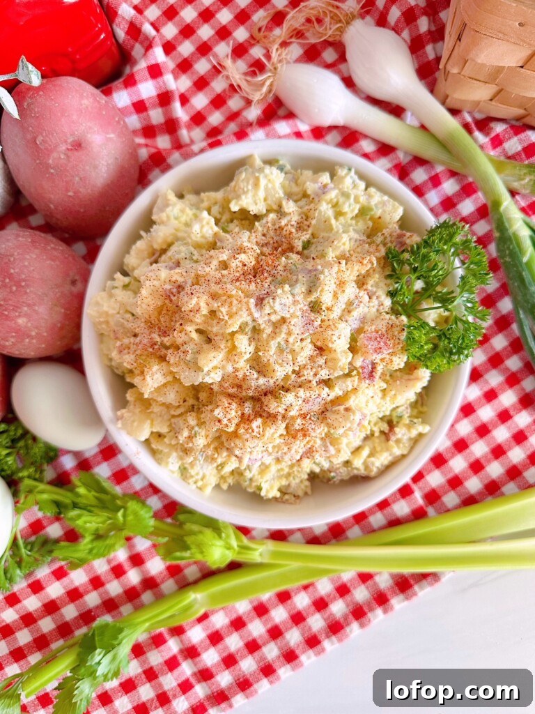 Classic Red Potato Salad with all the ingredients surrounding it in a white serving bowl on a red and white checkered cloth.