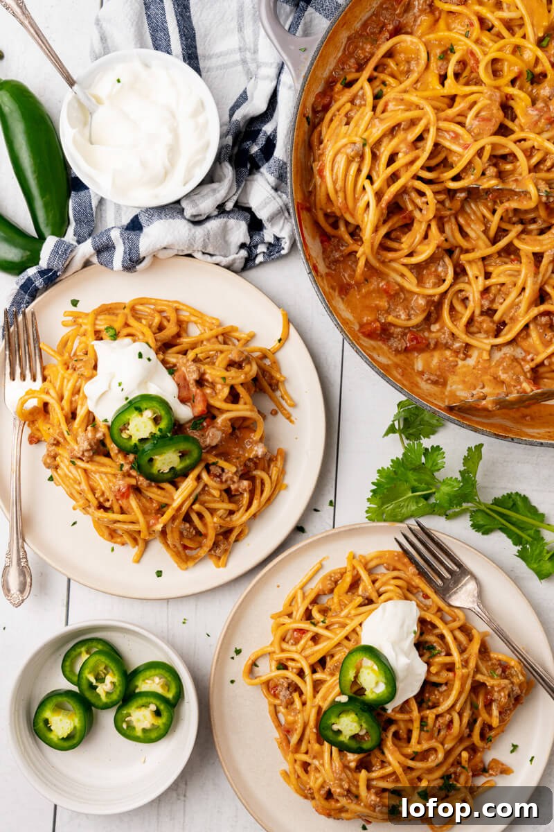 Two servings of Cheesy Taco Spaghetti with all the toppings on two white plates next to the pot of cheesy taco spaghetti.