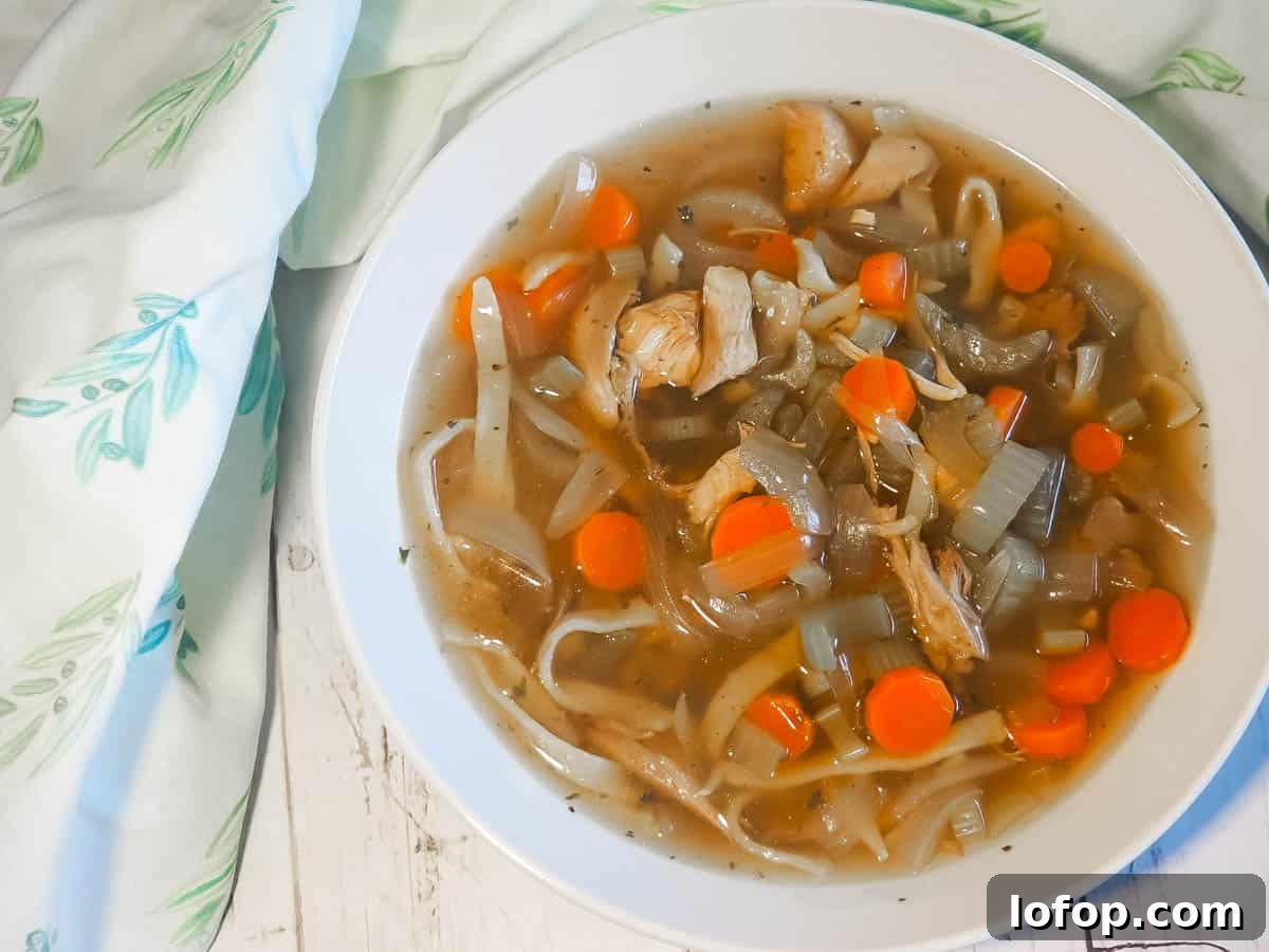 Paleo Chicken Noodle Soup with Homemade Egg Noodles Recipe 2 A white bowl of paleo chicken noodle soup with carrots, celery, and onions on a white wooden background with a blue and white tea towel.