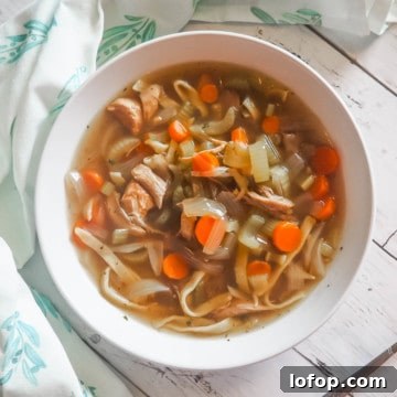 Paleo Chicken Noodle Soup with Homemade Egg Noodles Recipe 7 A white bowl of paleo chicken noodle soup with carrots, celery, and onions on a white wooden background with a spoon and blue and white tea towel.