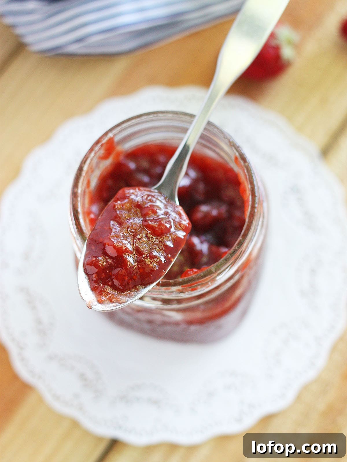 Close up shot of a glass jar of strawberry fig preserves on a white doily with a metal spoon of preserves resting on the top.
