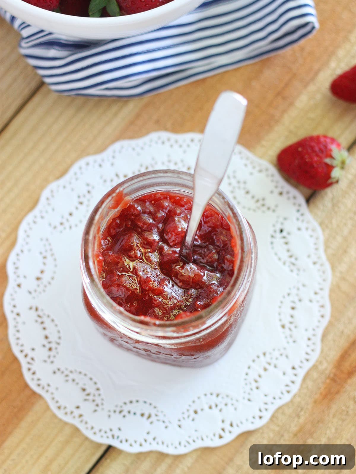 Glass jar of strawberry fig preserves on a white doily with a metal spoon sticking out.