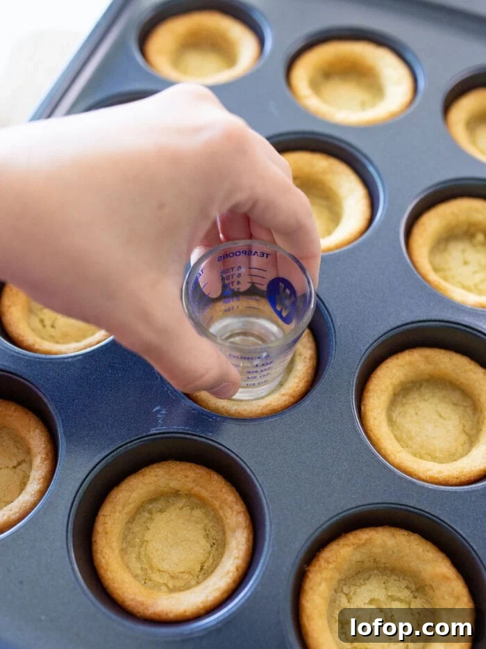 A hand presses a measuring cup into warm cookie dough cups in a muffin tin, creating indentations. The cookies are golden brown and evenly baked, with the tins compartments clearly visible.