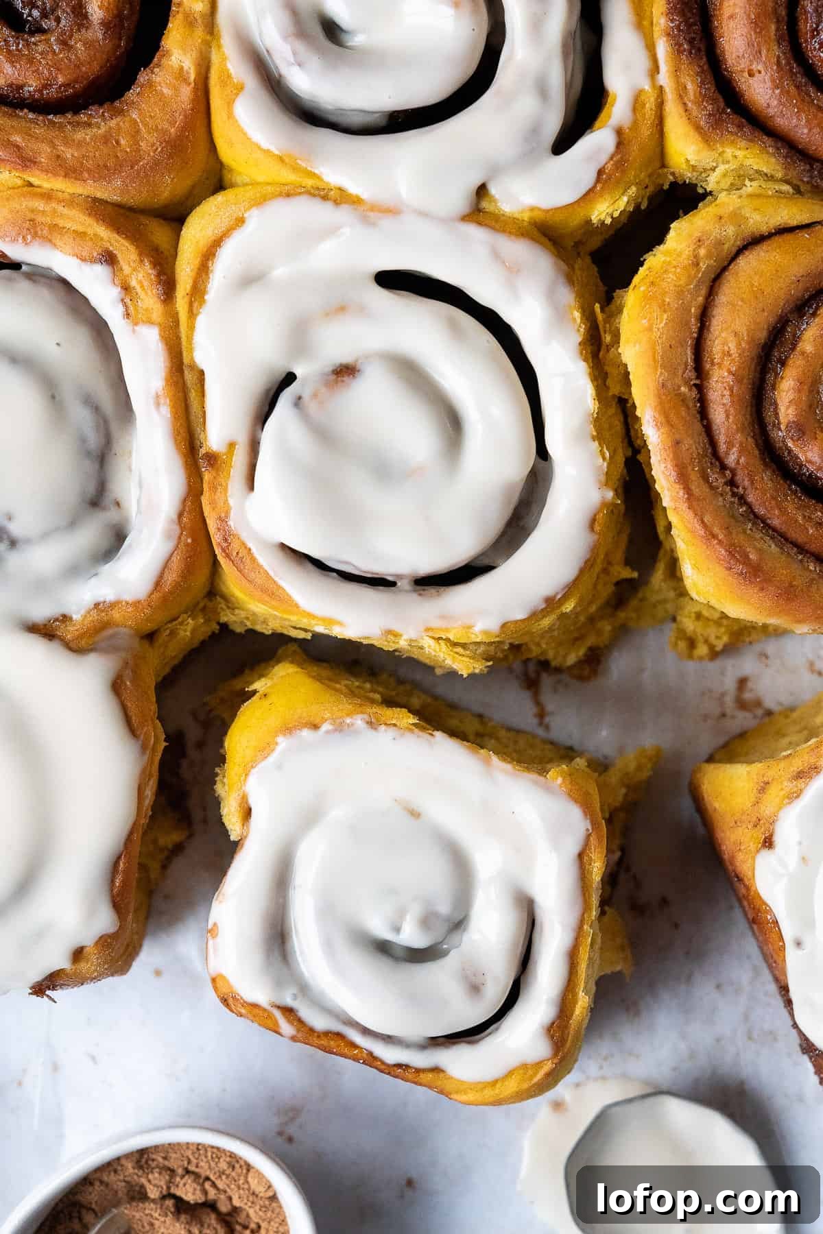 Vegan pumpkin cinnamon rolls on white baking parchment with a spoon of maple glaze and a bowl of pumpkin spice.