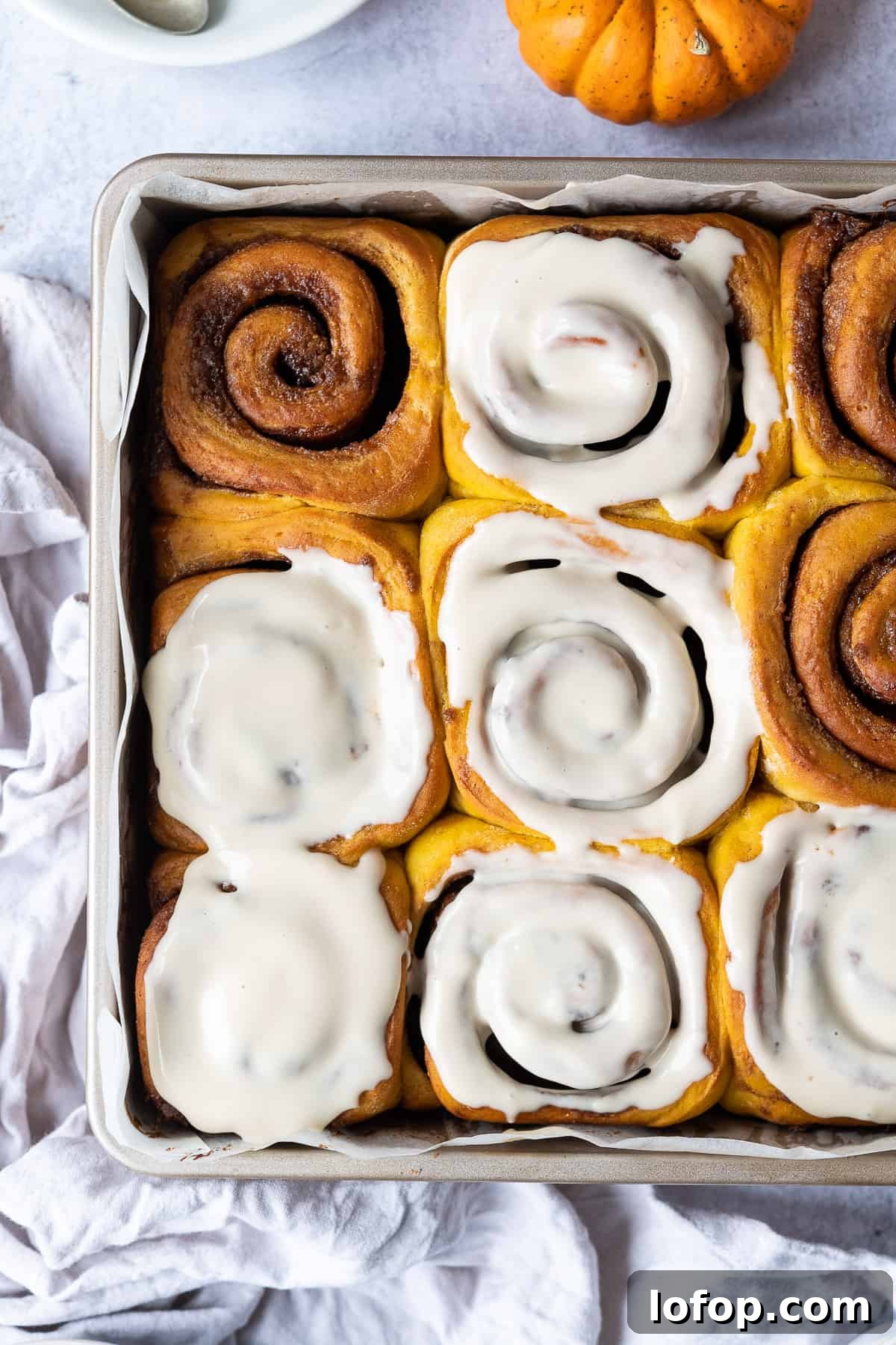 Vegan pumpkin cinnamon rolls with maple cream cheese glaze in a metal cake tin with a grey cloth and mini pumpkin.