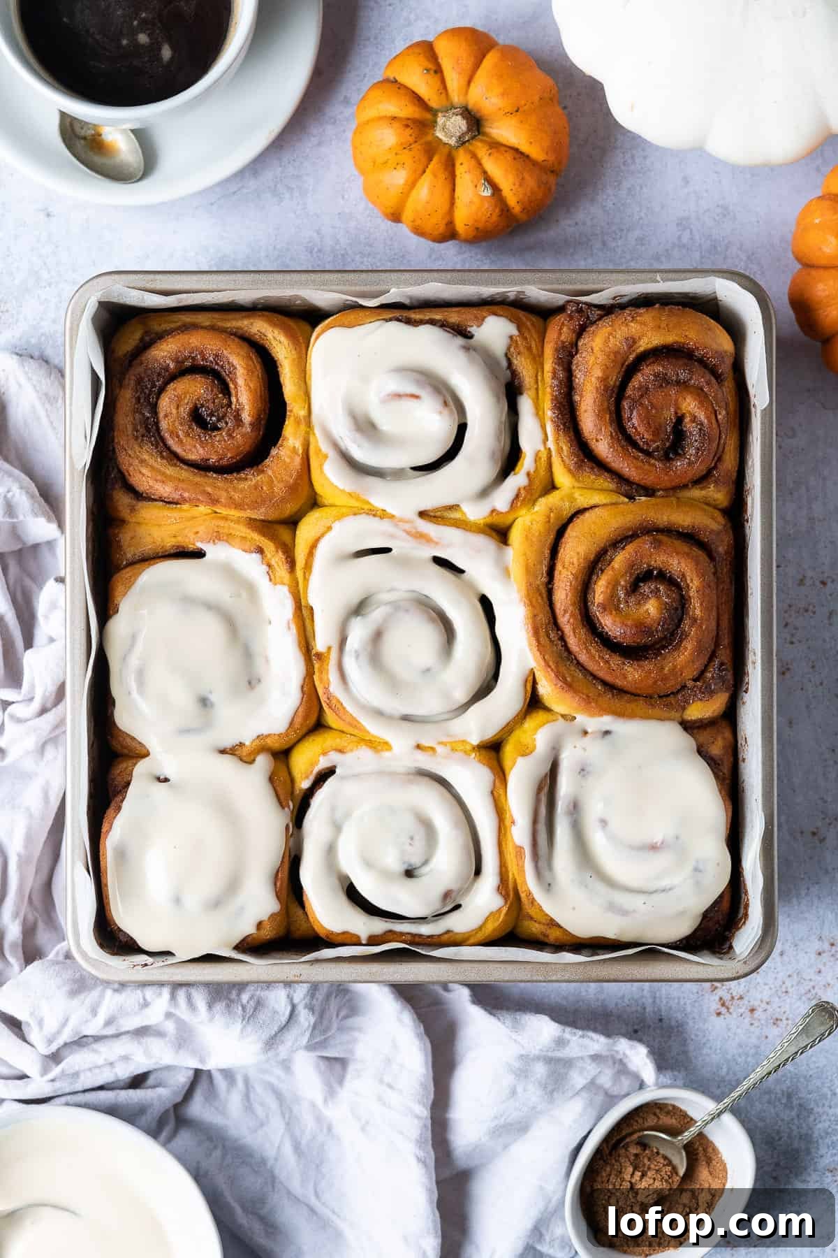 Vegan pumpkin cinnamon rolls in a metal baking tin on a grey background with mini pumpkins and a cup of coffee.