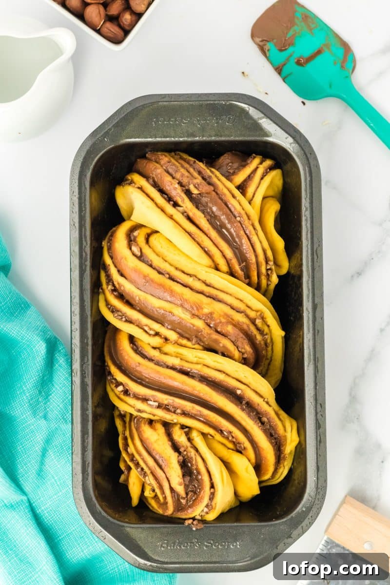 Top down view of a twisted pumpkin bread dough filled with nutella and chopped hazelnuts in a loaf pan - Hostess At Heart