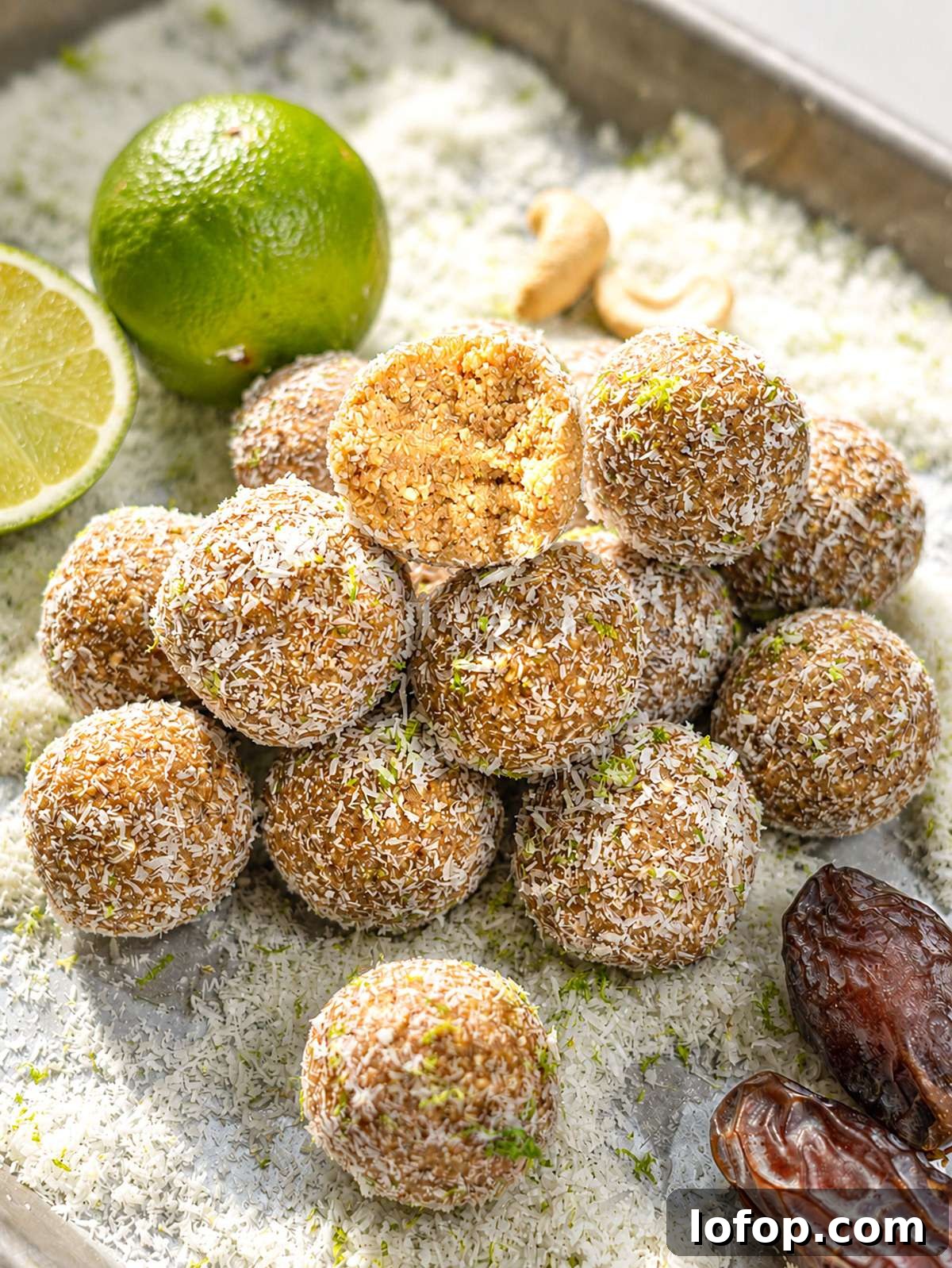 Coconut-covered key lime energy balls on marble with fresh limes, dates, and cashews nearby.
