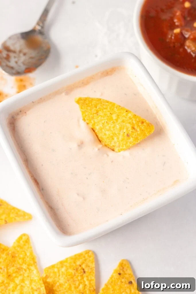 Close up of a white square dish with a salsa sour cream dip in it and a yellow tortilla chip sticking out of it. There is salsa, a spoon, and other tortilla chips surrounding the dish.