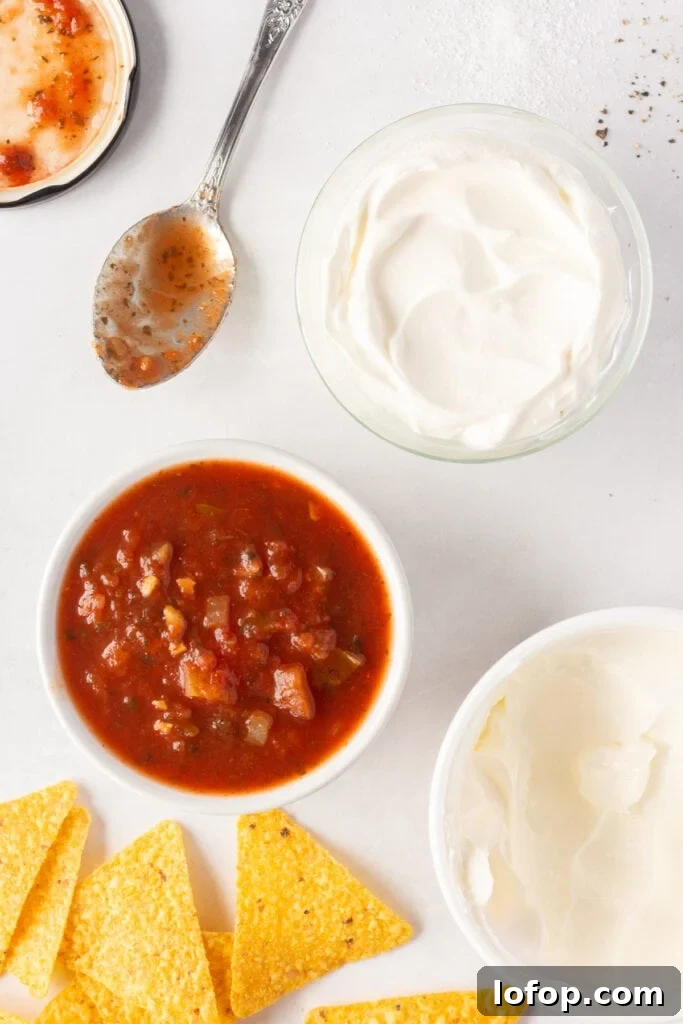 Top down shot of two bowls, one with salsa the other with sour cream, on a white background. They're surrounded by a spoon with salsa on it. a container of sour cream, and yellow tortilla chips.