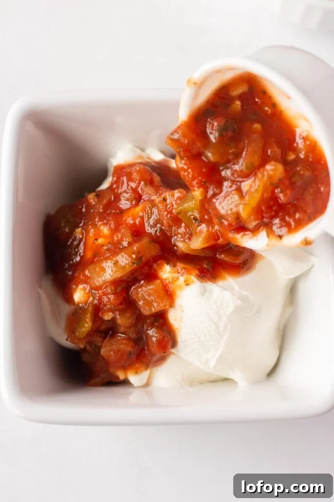 Red salsa being poured out of a measuring cup into a white square dish with sour cream in it.