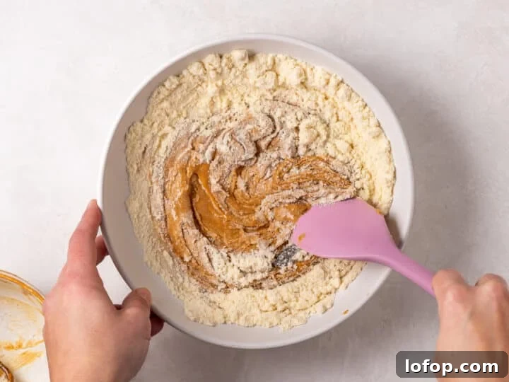 A pink spatula stirring peanut butter, protein powder, vanilla bean paste, and pure maple syrup together in a mixing bowl