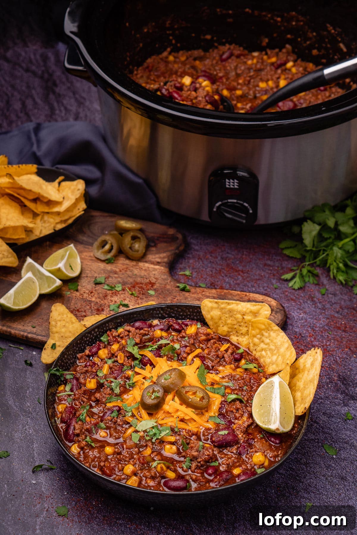 Slow Cooker Chilli Con Carne in a black bowl with red leicester cheese, jalapenos, wedge of lime and tortilla chips served on top and a bowl of tortilla chips and a slow cooker filled up with chilli in the background
