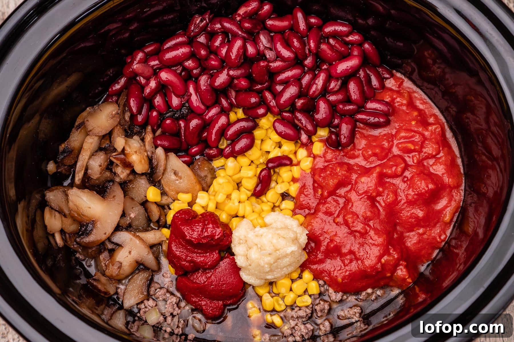 A slow cooker pot with mince beef, chopped onion, sweetcorn, chopped tomatoes garlic, tomato puree, mushrooms and kidney beans inside