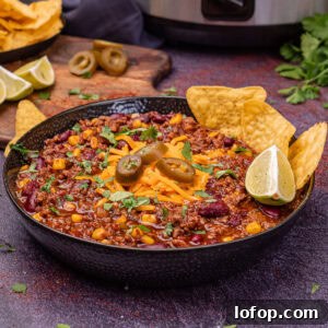 Slow Cooker Chilli Con Carne in a black bowl with red leicester cheese, jalapenos, wedge of lime and tortilla chips served on top