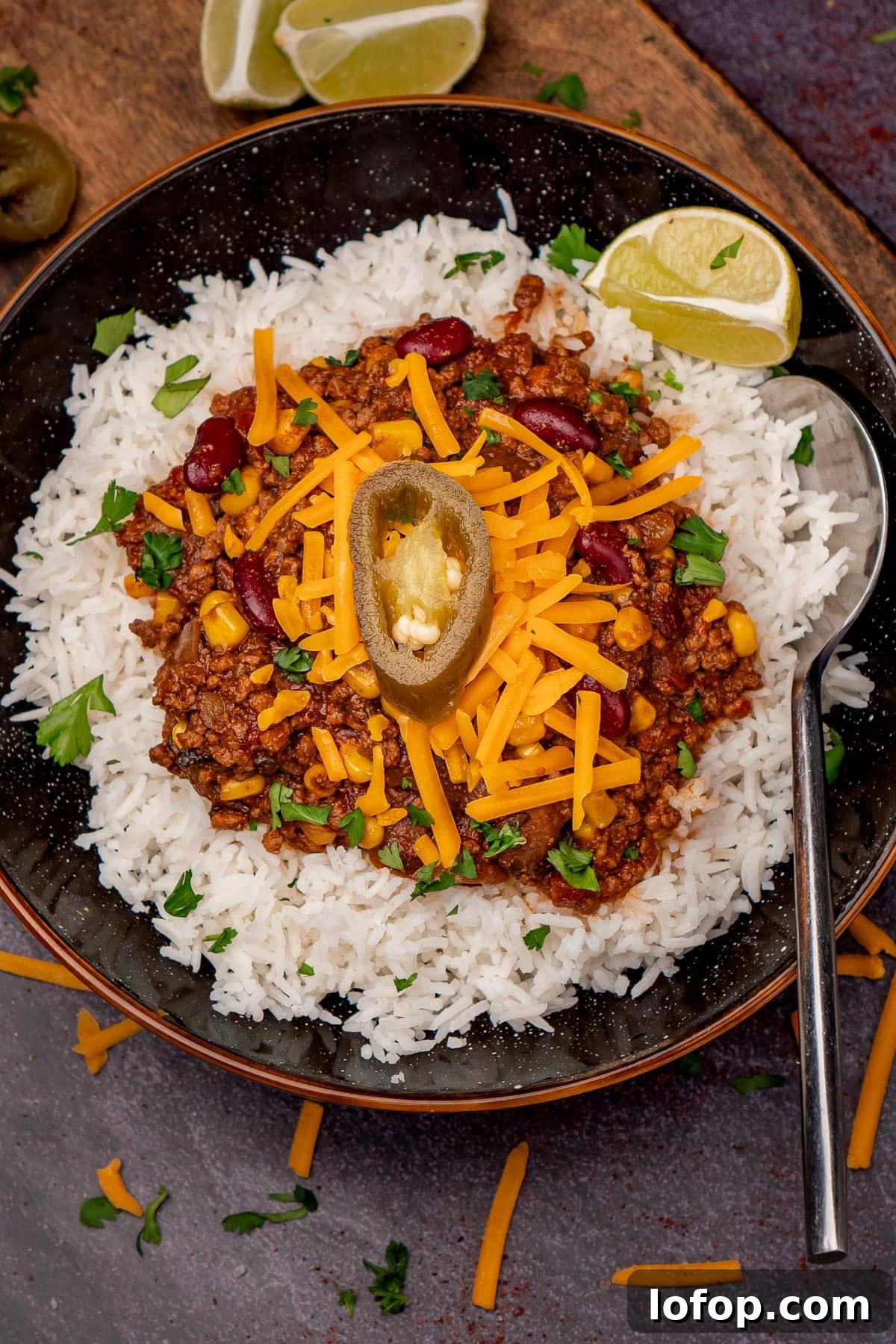 Slow Cooker Chilli Con Carne served with white rice and red leicester cheese and a jalapeno on top and a silver spoon with a wedge of lime on the side