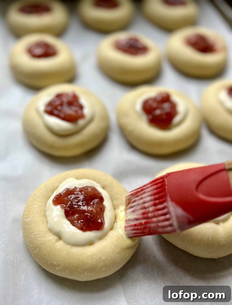 Brushing egg wash on sourdough kolaches filled with cream cheese filling and fruit jam.