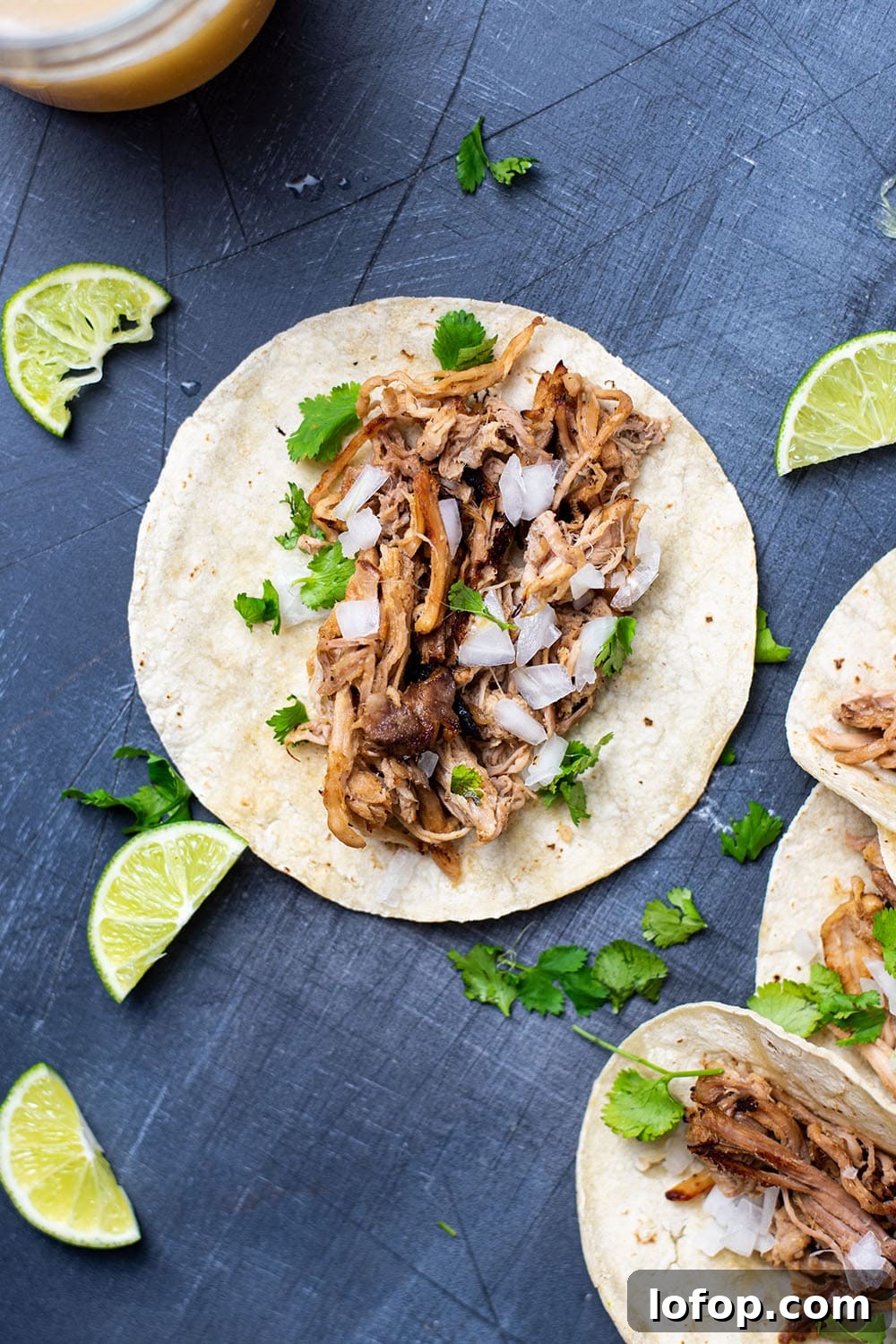 Slow Cooker Pork Carnitas Tacos Recipe for Tender Flavor 3 A pork carnita taco topped with white onion and cilantro on a dark surface next to lime wedges.