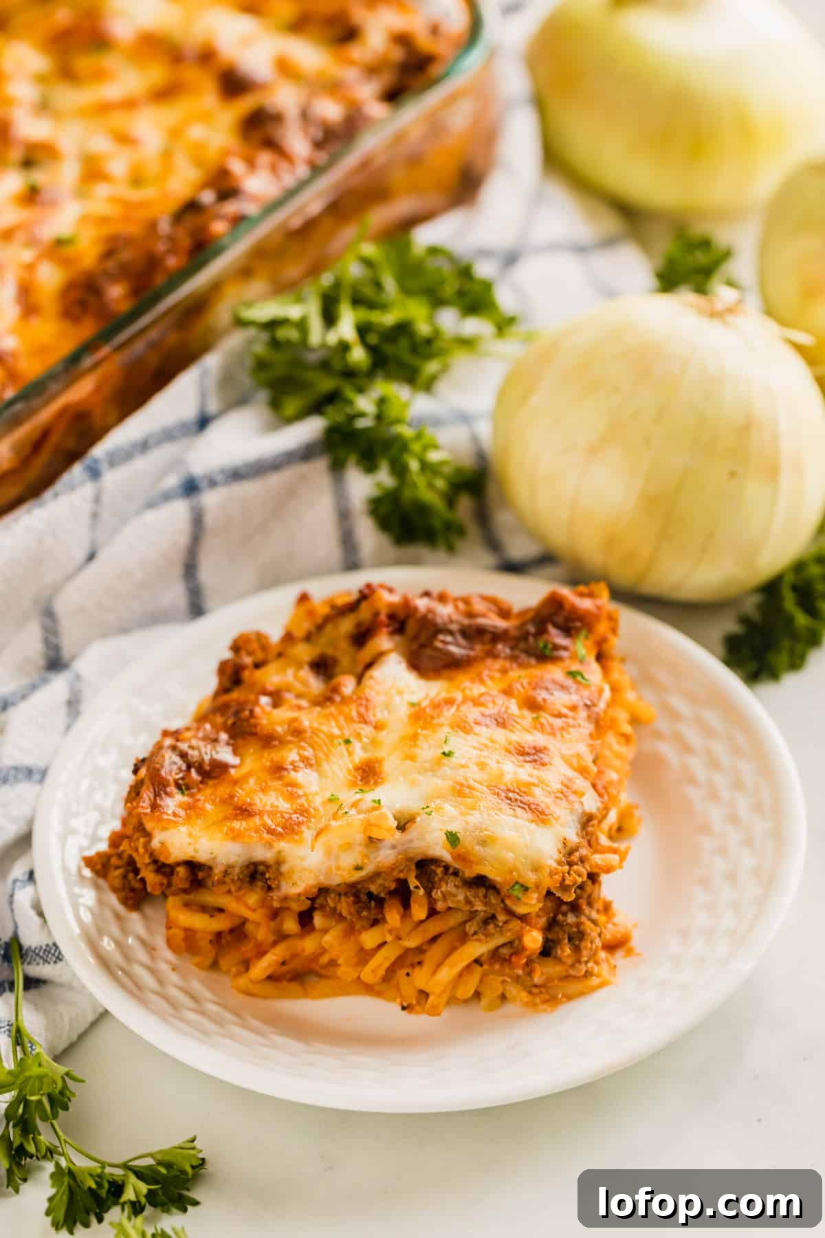 Ultimate Cheesy Baked Spaghetti Recipe for Comfort Dinner 11 A piece of baked spaghetti casserole on a plate with onions, herbs and the rest of the pan behind it.