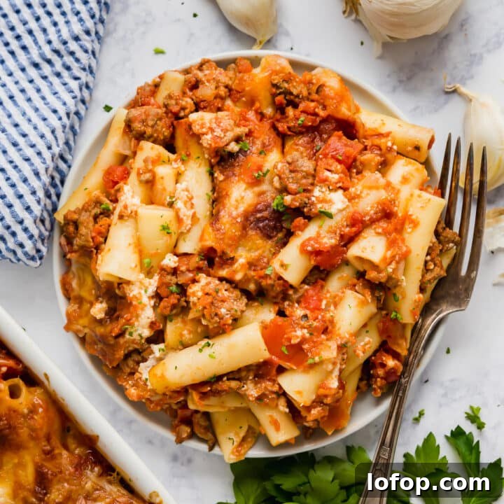 An overhead image of a plate of baked ziti with herbs, garlic and a blue striped napkin next to it.