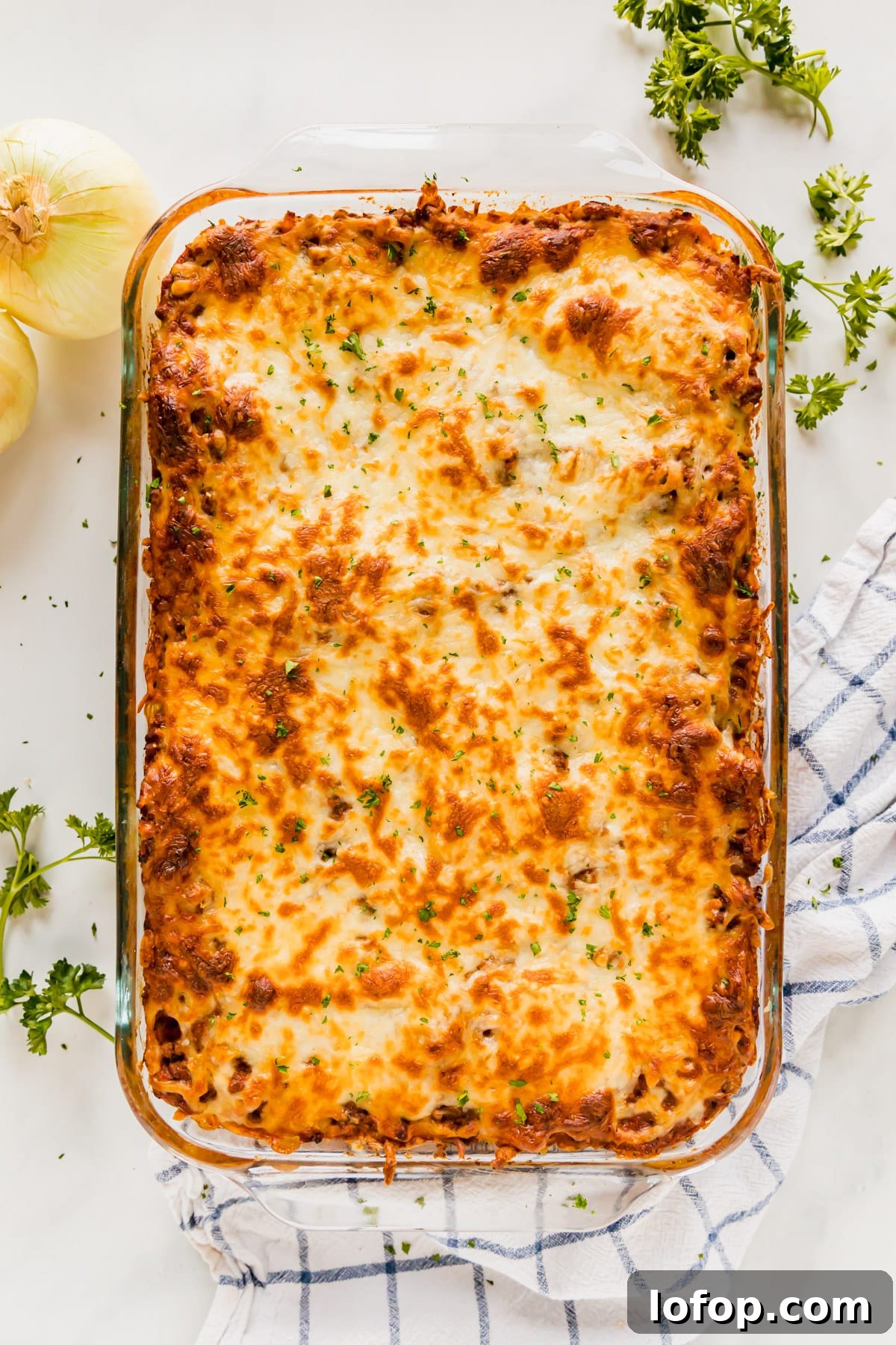 Ultimate Cheesy Baked Spaghetti Recipe for Comfort Dinner 3 An overhead image of a pan of baked spaghetti with a towel, onions and herbs around it.