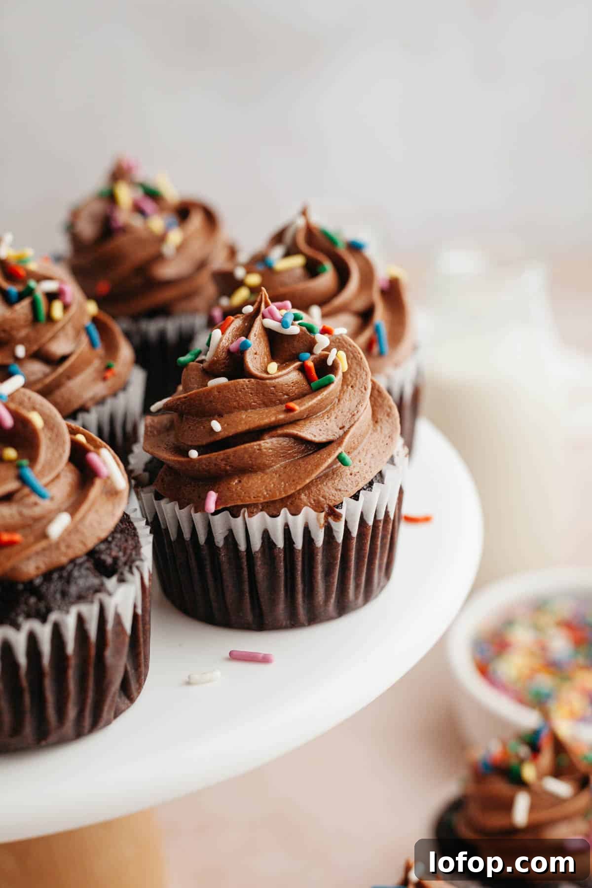 Decadent Chocolate Cupcakes with Cream Cheese Frosting 11 Chocolate cupcakes with chocolate frosting and rainbow sprinkles on a white cake stand.