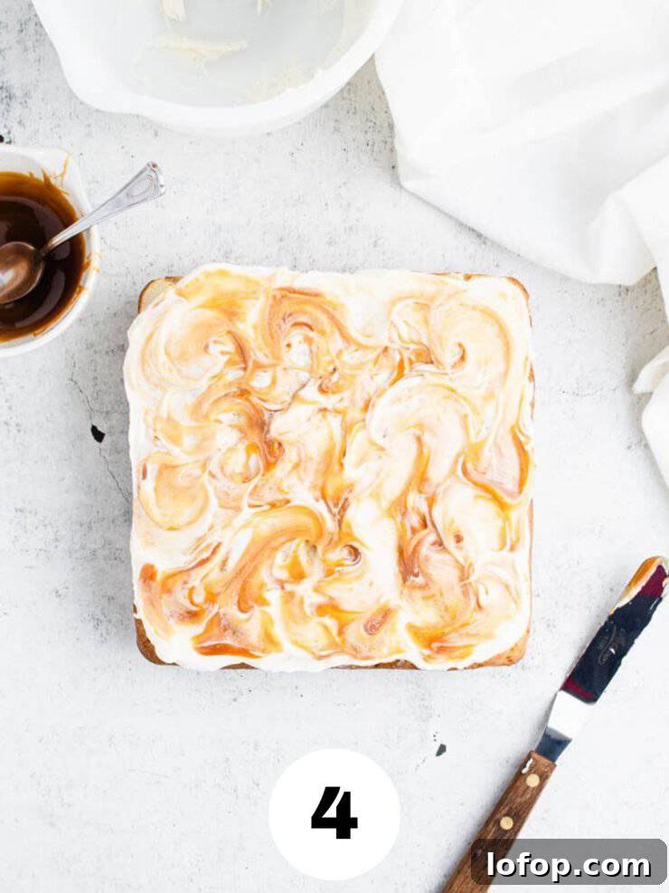 brown butter frosted apple cake with swirls of caramel next to a bowl of caramel sauce and an offset spatula with a wooden handle.