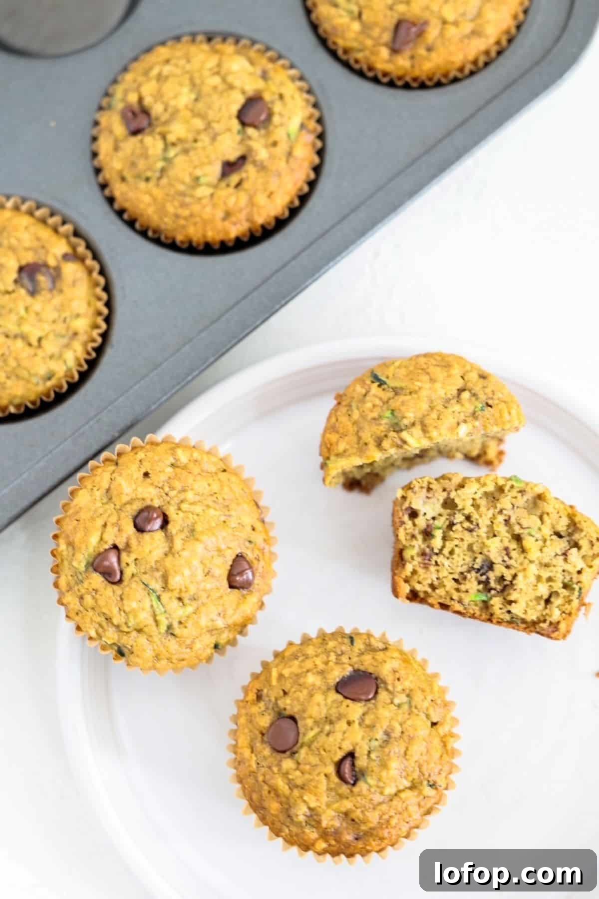 Two whole and two halves of banana chocolate chip muffins  on a whit plate with a muffin tin of muffins behind it.