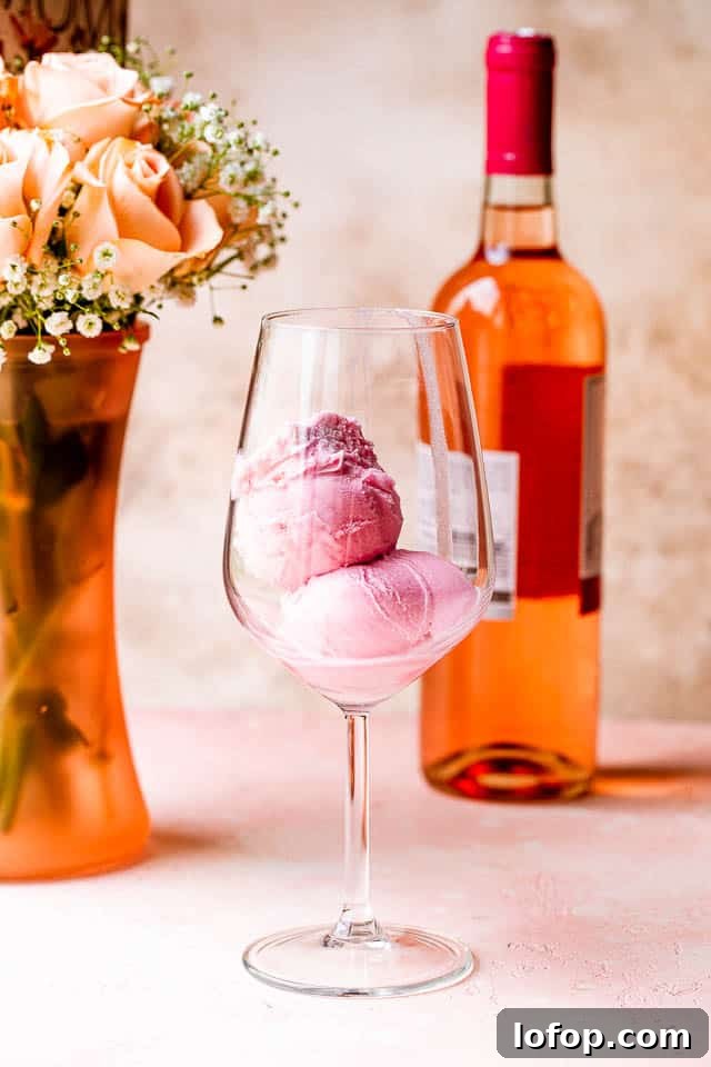 Two scoops of sorbet in a wine glass in front of a vase of roses and a bottle of rosé.