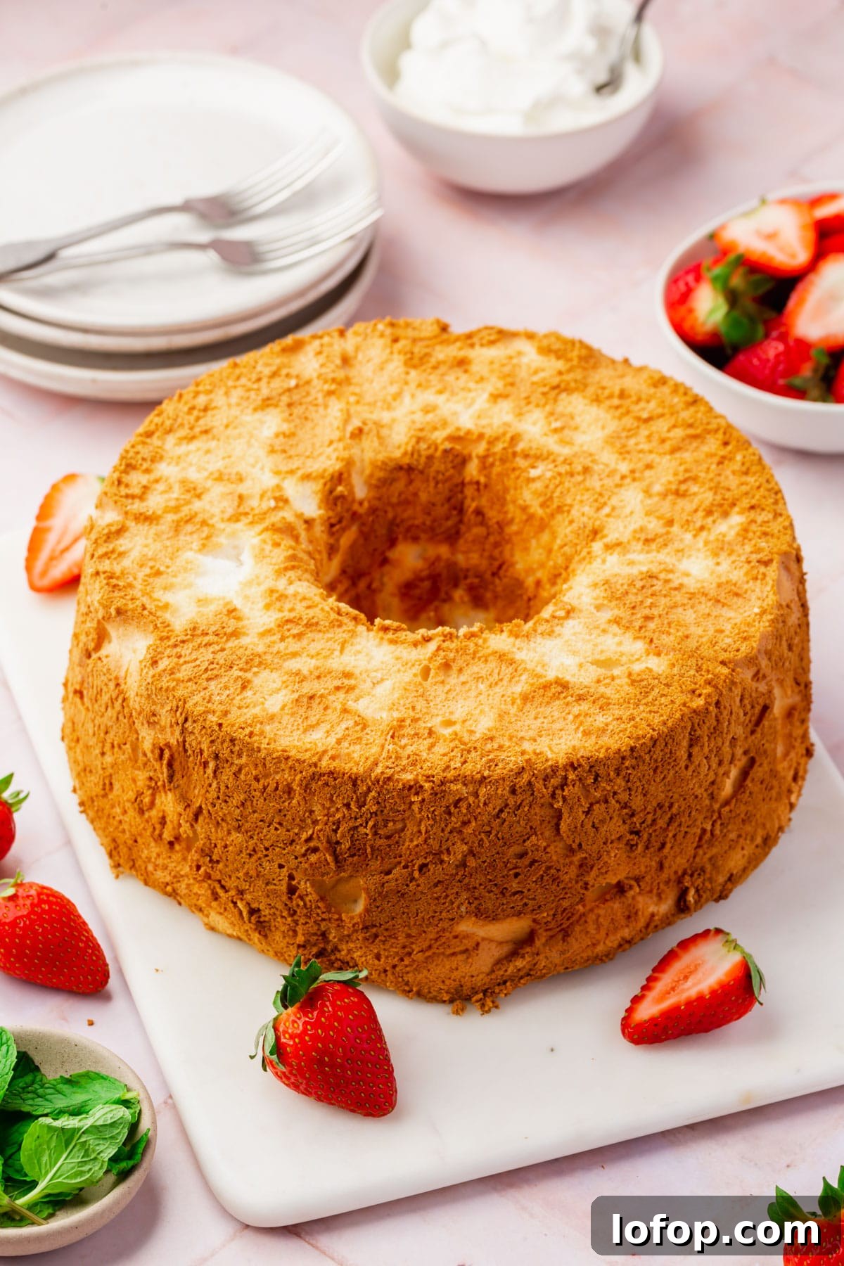 Fluffy Gluten-Free Angel Food Cake Recipe for Light Desserts 2 An angel food cake on a serving platter with strawberries scattered around, plates and forks in the background and small bowls of whipped topping, strawberries and mint leaves.