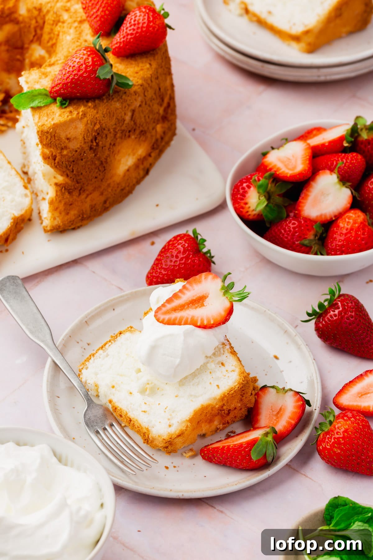 Fluffy Gluten-Free Angel Food Cake Recipe for Light Desserts 5 A slice of angel food cake on a plate topped with whipped cream and a strawberry with a full angel food cake and a bowl of strawberries in the background.