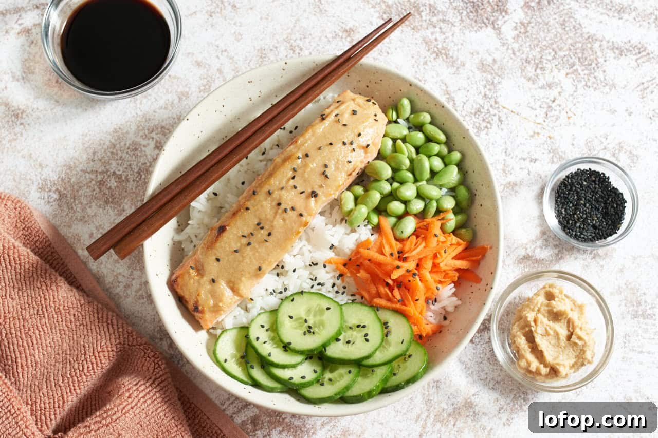 Miso Butter Salmon Rice Bowl Recipe 2 Miso butter salmon in a bowl with rice, edamame, carrots, and cucumber, topped with black sesame seeds, small bowls of condiments surround it, chopsticks rest on the left side of the bowl.