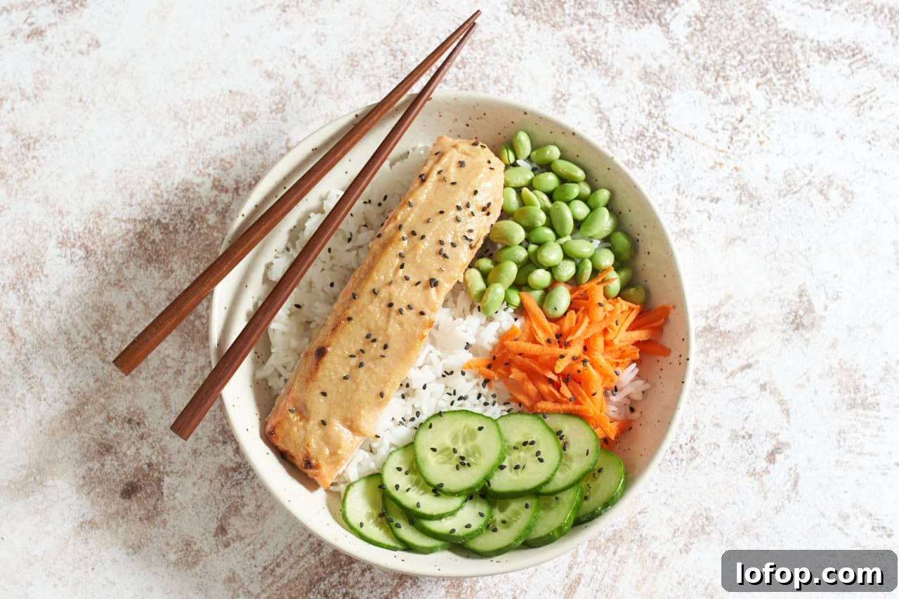 Miso Butter Salmon Rice Bowl Recipe 6 Miso butter salmon in a bowl with rice, edamame, carrots, and cucumber, topped with black sesame seeds, with chopsticks resting on the left side of the bowl.