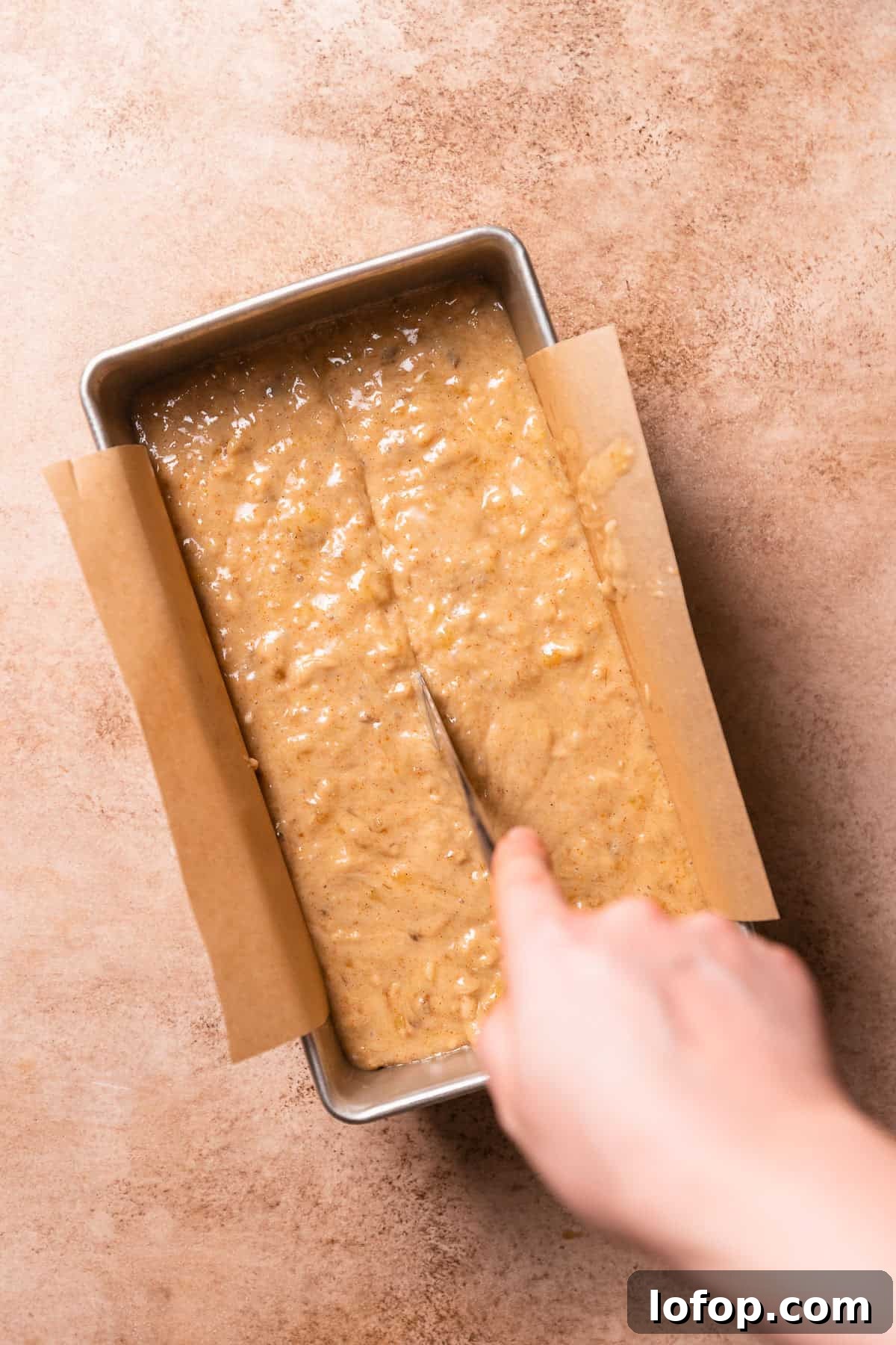 A hand dragging a butter knife through the center of the batter in a loaf pan.