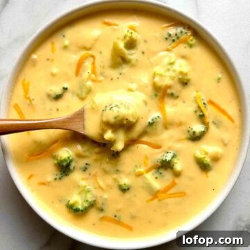 overhead shot of broccoli cheddar soup in white bowl; wooden spoon lifting up some of the soup.