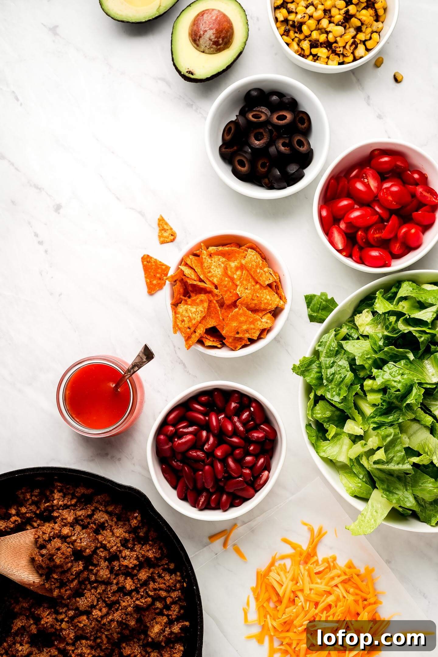 Crispy Dorito Taco Salad Recipe for Game Day and Weeknight Meals 5 Ingredients on a marble surface- roasted corn, avocado, black olives, tomatoes, Doritos, lettuce, kidney beans, shredded cheese, taco meat, and dressing.