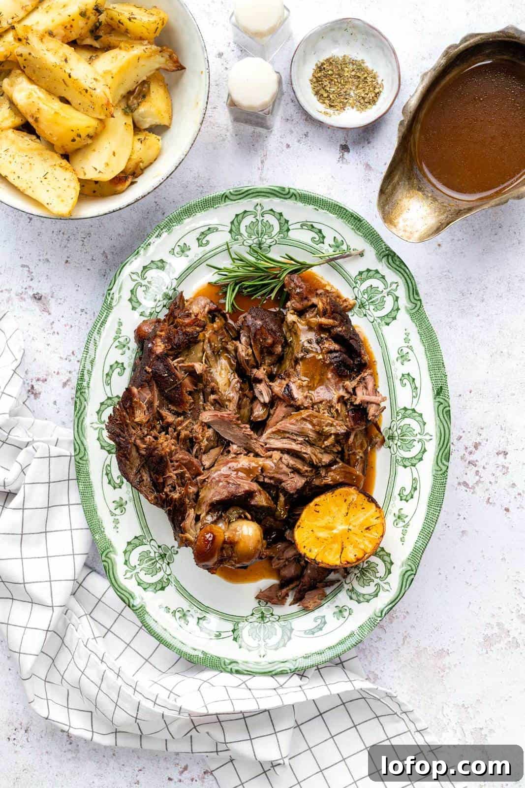 Slow Cooker Leg of Lamb with Garlic and Herbs 2 A green-rimmed platter with shredded Slow Cooker Lamb, roasted lemon, and rosemary sprigs, surrounded by a bowl of herbed potato wedges, gravy boat, seasonings, and a white checkered cloth.