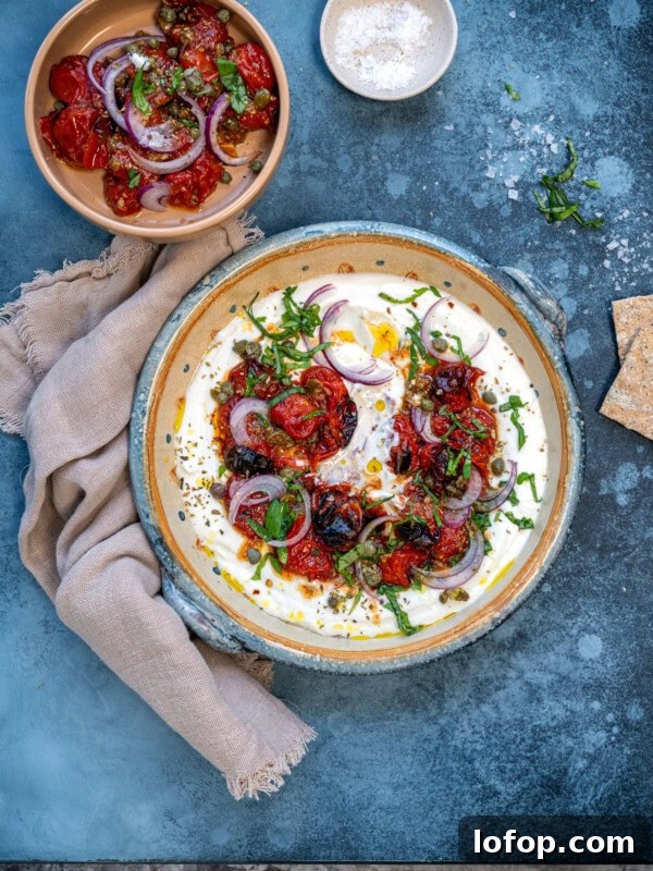 Slow Cooker Leg of Lamb with Garlic and Herbs 11 A dish on a blue surface features roasted tomatoes, red onions, and herbs on whipped cottage cheese dip. A smaller dish of tomatoes and onions is beside it. A light-colored napkin, a small bowl of salt, and cracker pieces are also present.