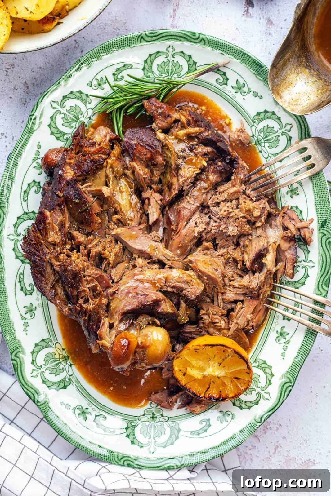 Slow Cooker Leg of Lamb with Garlic and Herbs 3 Shredded, juicy slow cooker lamb with gravy on a green-patterned platter, garnished with fresh rosemary and a grilled lemon half; a fork rests on the side of the meat.