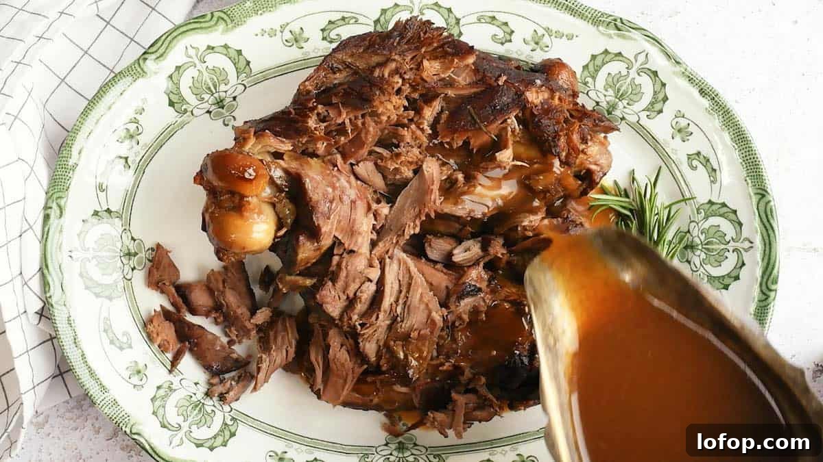 Slow Cooker Leg of Lamb with Garlic and Herbs 4 A platter of Slow Cooker Lamb, shredded with the bone, is served on a decorative green and white plate as brown gravy is poured over the tender meat. A sprig of rosemary sits on the side for garnish.
