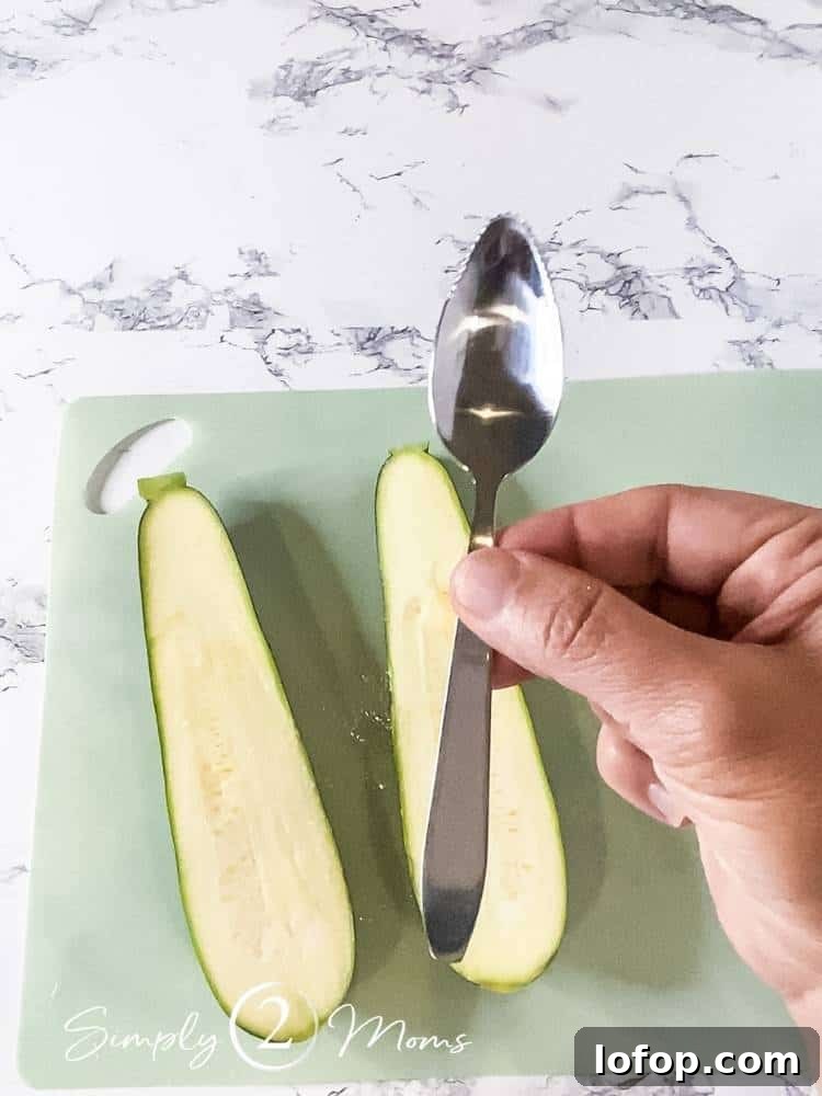 A hand holding a serrated grapefruit spoon above a fresh zucchini that's been cut in half on a cutting mat