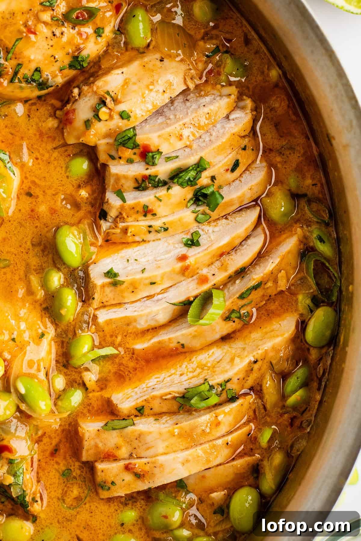 Close-up of sliced cooked chicken in a pan with a creamy, orange-colored sauce, garnished with chopped herbs and green peas.