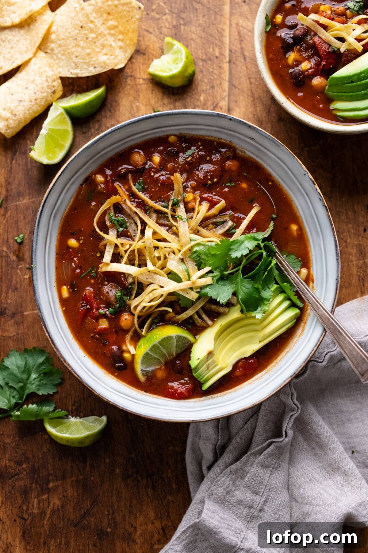 Hearty Vegetarian Black Bean Tortilla Soup Recipe 2 Ceramic bowls with vegetarian tortilla soup in them sitting on a wooden background. On top of the soup is strips of tortillas, avocado slices, and cilantro, with lime slices and a gray napkin arranged around the bowls.