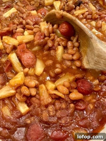 Baked beans, franks and pineapple pieces in a baking dish with a spoon.
