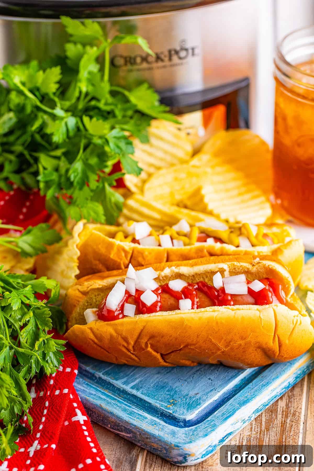 Easy Crockpot Hot Dogs Recipe served on a fluffy bun with toppings in front of a black slow cooker filled with cooked hot dogs.