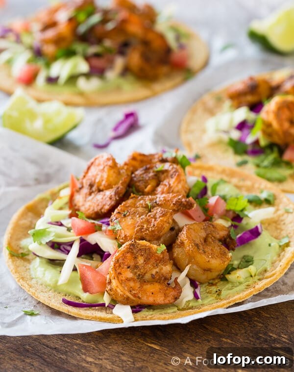 Baja Shrimp Tacos Recipe — Grilled or Stovetop Method 2 Grilled shrimp tacos piled on white paper on a wood table, with avocado sauce and veggies.