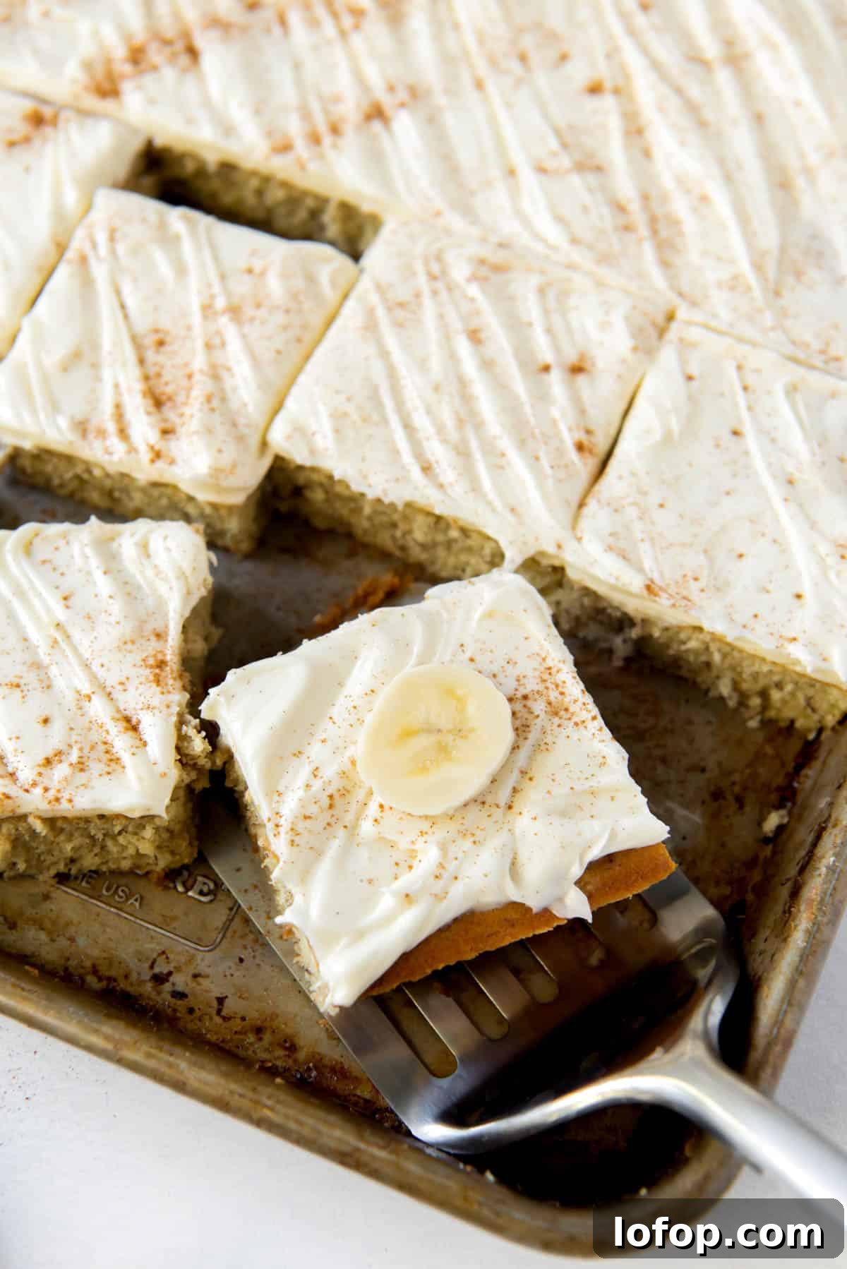 Banana bars with cream cheese on a sheet pan, one slice being served.