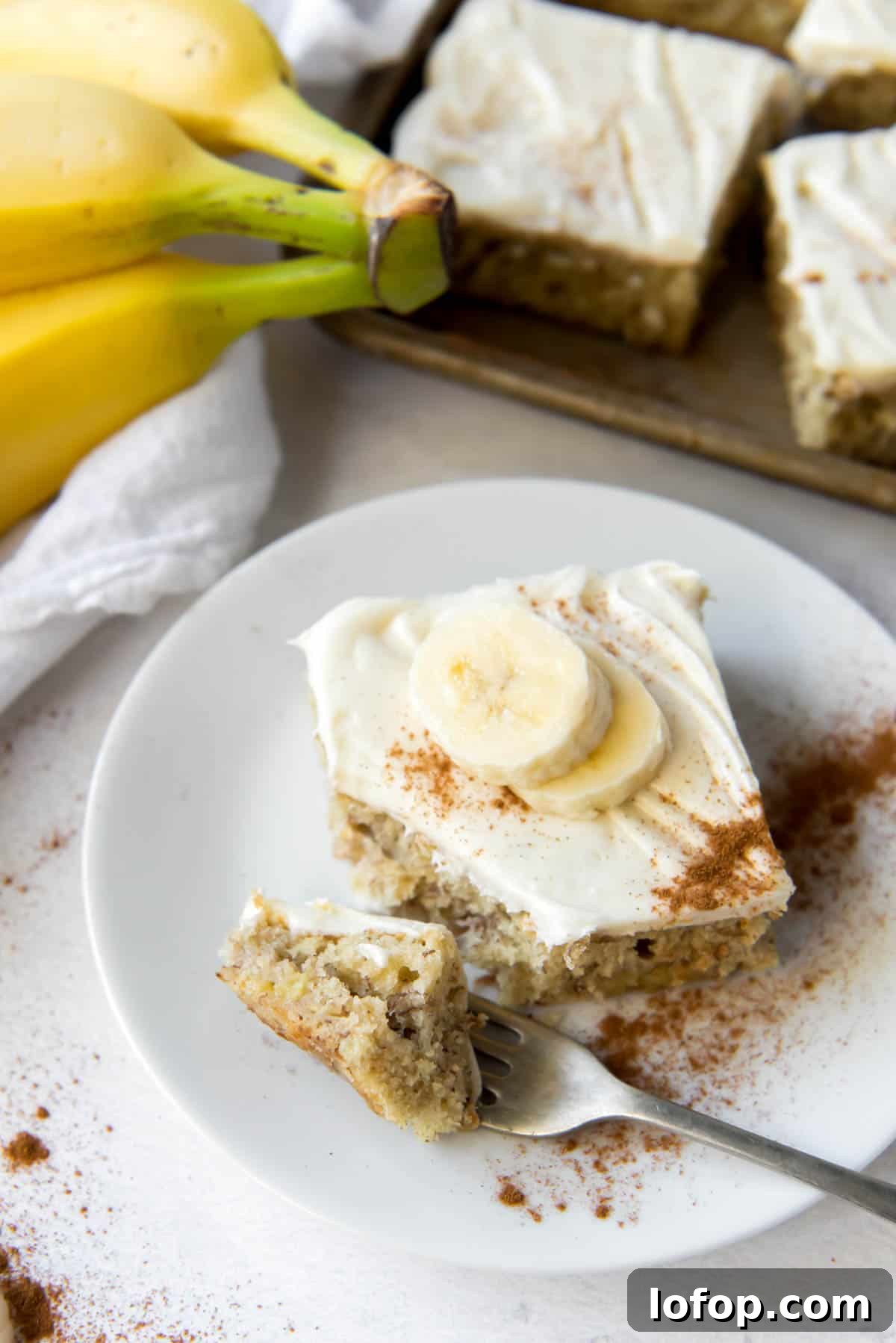 A banana bar with cream cheese icing on a plate, with bananas and more bars in the background.