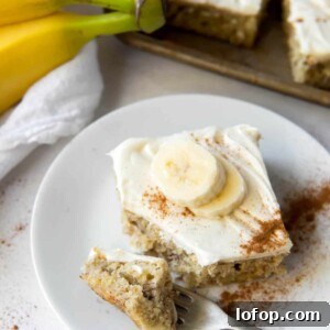 A banana bar with cream cheese icing on a plate, with bananas and more bars in the background.