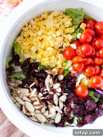 White bowl containing corn, cherry tomatoes, lettuce, almonds, and cranberries.